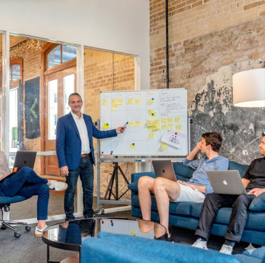 Mann präsentiert am Whiteboard, während drei Personen auf einer Couch mit Laptops arbeiten.